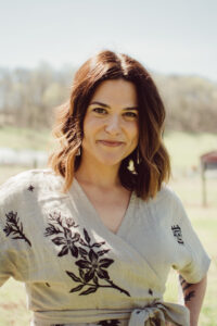 Claire Crunk_Headshot_2026 Smiling woman with shoulder-length brown hair, wearing a grey wrap dress with black floral patterns, outdoors on a sunny day.