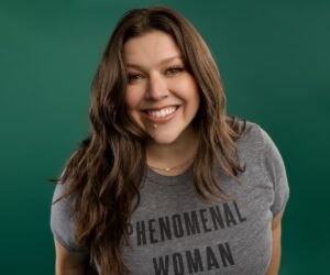 Smiling woman with long brown hair wearing a gray shirt that reads "PHENOMENAL WOMAN," against a vibrant green background.