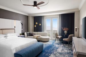 Luxurious hotel room featuring a king bed, a curved sofa, stylish decor, and large windows with a view of the outdoors.