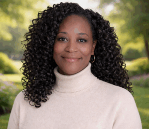 deidre goodwin Smiling woman with curly black hair, wearing a beige turtleneck, against a blurred green and floral garden background.