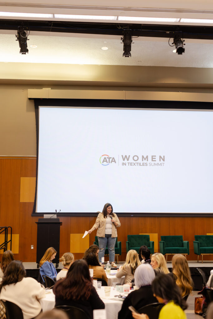 A speaker stands on stage at the Women in Textiles Summit, addressing an audience seated at round tables with green chairs in the background.