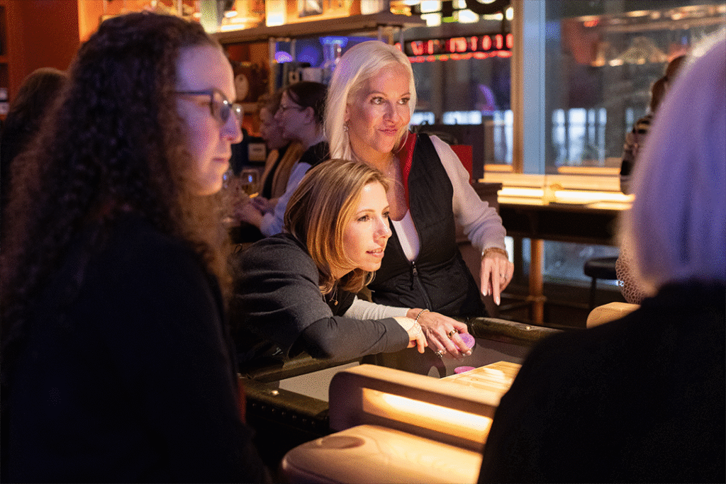 Three women engage around a low table in a dimly lit space, with warm tones and glowing light features enhancing the lively atmosphere.