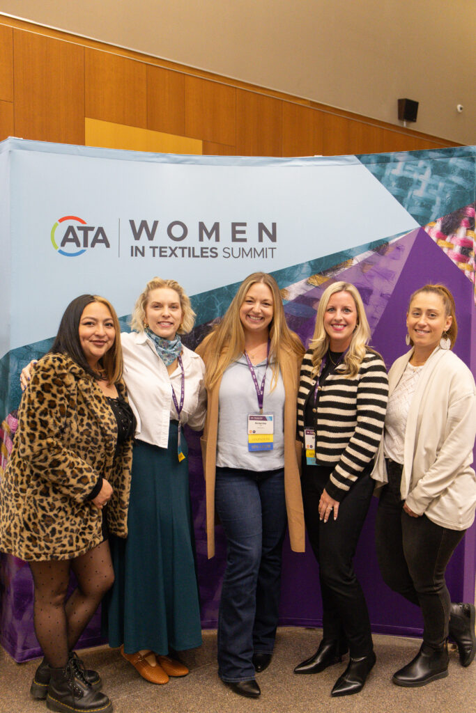 Five women pose in front of a colorful backdrop for the Women in Textiles Summit, showcasing diverse styles and smiles.