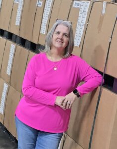 Donnapic A woman in a bright pink long-sleeve shirt stands confidently, leaning against a row of cardboard boxes in a warehouse setting.