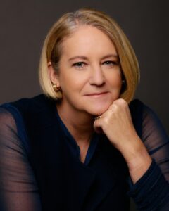 JKnightHeadshot Blonde woman with chin resting on her hand, wearing a dark vest over a blue shirt, set against a dark grey background.