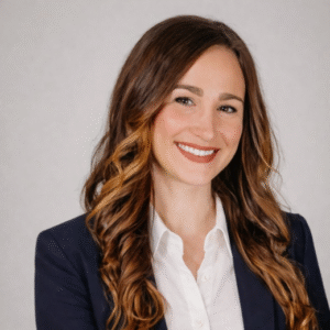 Jenny Nichols Smiling woman with long, wavy hair wearing a navy blazer and white shirt, set against a light gray background.