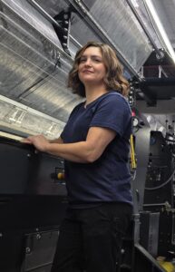 Tonya pic Woman with shoulder-length brown hair stands confidently by a large machine, wearing a navy shirt and black pants, in a workshop setting.