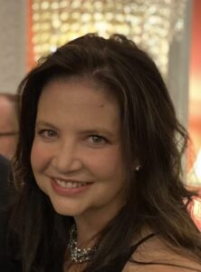 amy jenkins A woman with long, dark hair smiles warmly, wearing a sparkling necklace, against a softly blurred background of warm colors.