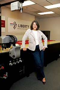 A woman in a light jacket stands beside equipment in a workspace featuring a "Liberty" banner, with a modern, professional atmosphere.