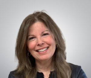 Screenshot Smiling woman with long, brown hair and hoop earrings, wearing a black top, set against a soft gray background.
