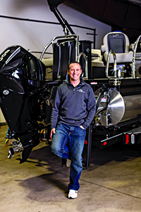 A man in a grey sweatshirt and jeans stands confidently beside a sleek black boat motor, set against a spacious garage backdrop.