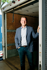 0073_zach_wilson_greenwich_tent_company_photojane-Edit_pxl A man in a blazer and checkered shirt stands by a truck's open door, smiling confidently against a wooden interior backdrop.