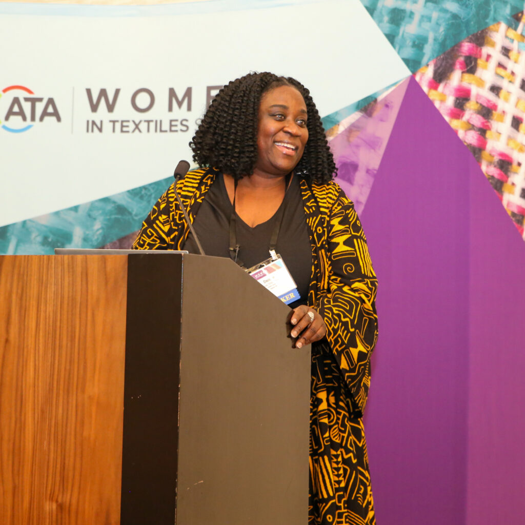 ata_womenintextiles26_190_55203538311_o A woman with curly hair wearing a patterned yellow and black wrap stands at a podium, smiling. A colorful backdrop features abstract shapes.
