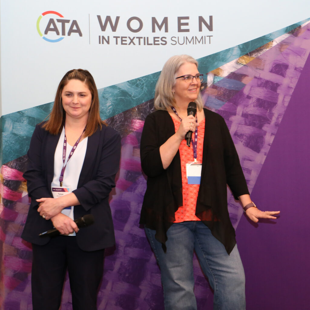 ata_womenintextiles26_215_55203931435_o Two women speaking at a conference. One wears a navy blazer and the other, a black cardigan over an orange top. Colorful textile backdrop.