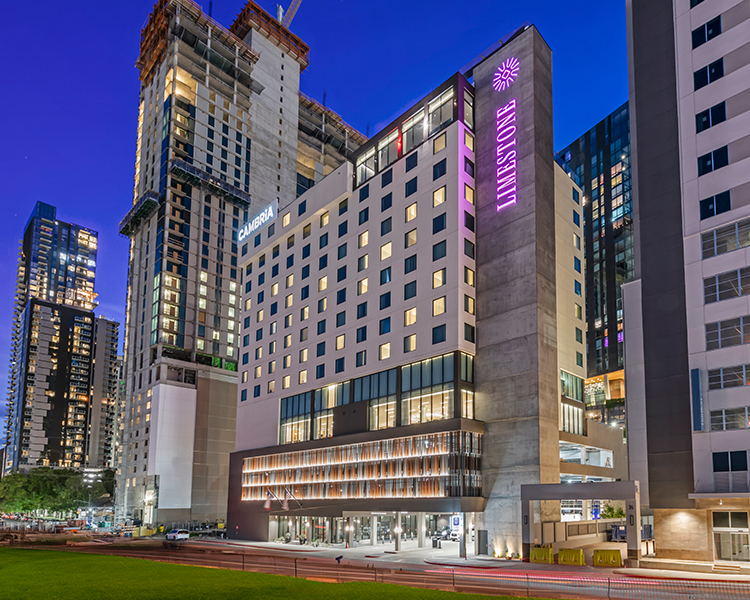 cambriahotelaustindowntown-homepage-staystepsfromraineystreet-657b682a0c205 Modern hotel building with large windows, featuring a lit sign. Surrounding construction and urban landscape under a twilight sky.